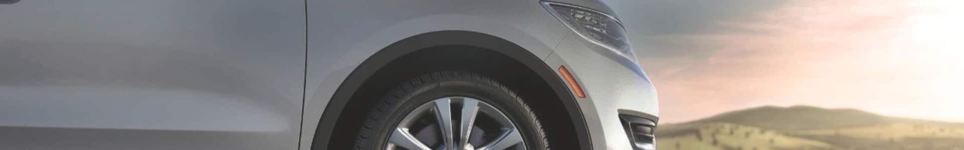 Front section of a silver car featuring a wheel and headlight, with a blurred landscape background.