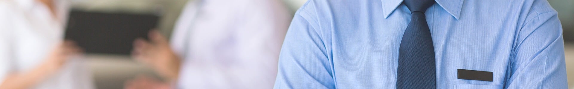 Person in blue shirt and tie, standing with arms crossed, with blurred figures in the background.