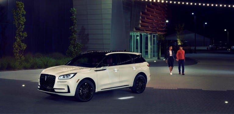 White SUV parked near a modern building at night, with two people walking nearby.